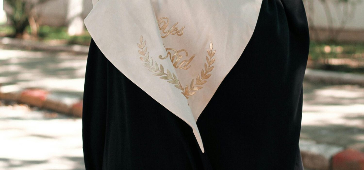 A woman wearing a hijab and graduation gown celebrates her achievement outdoors in Algiers.